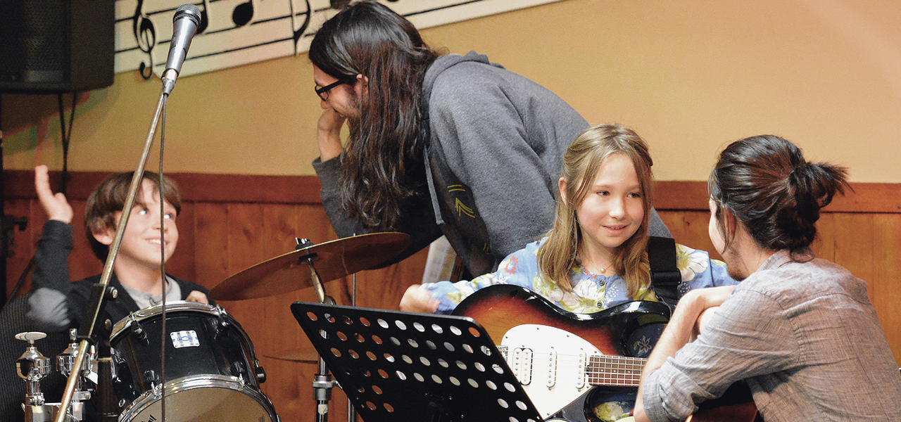Fille de 8-10 ans au cheveux roux jouant de la guitare électrique accompagné par sa professeure.