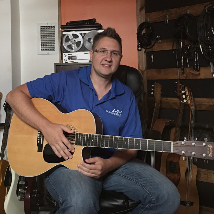 Jean-Pierre Lessard assis avec une guitare à la main et portant un chandail bleu avec le logo de Musique O Max.