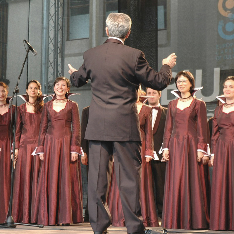 En avant-plan, on voit le chef de la chorale et en arrière-plan, on y voit plusieurs personnes qui chantent.